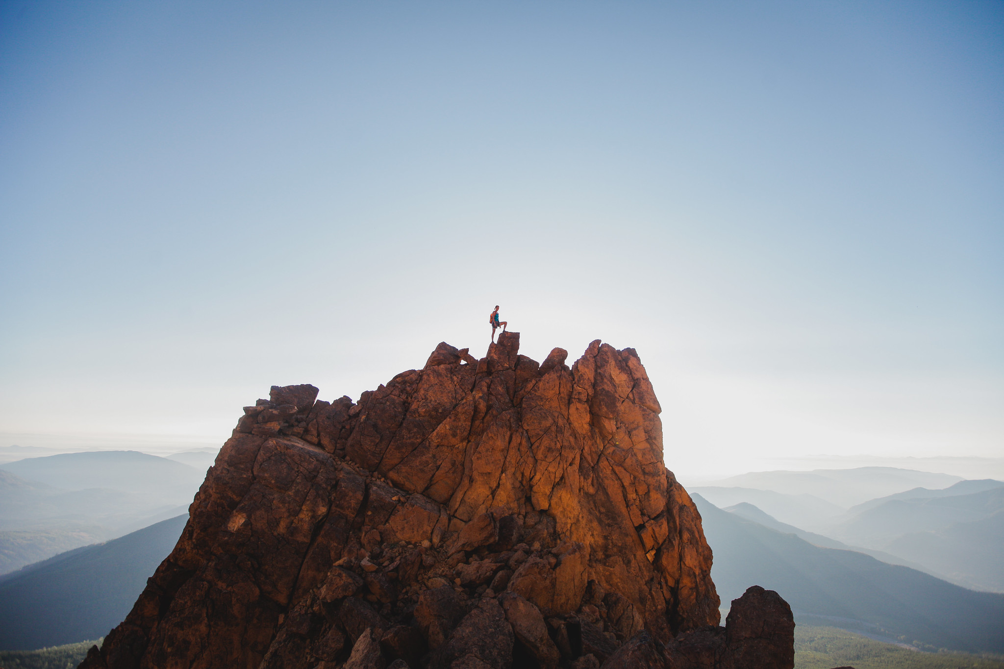 Person on the top of a tall mountain summit. In the background is an entire mountain range. The sky is clear.