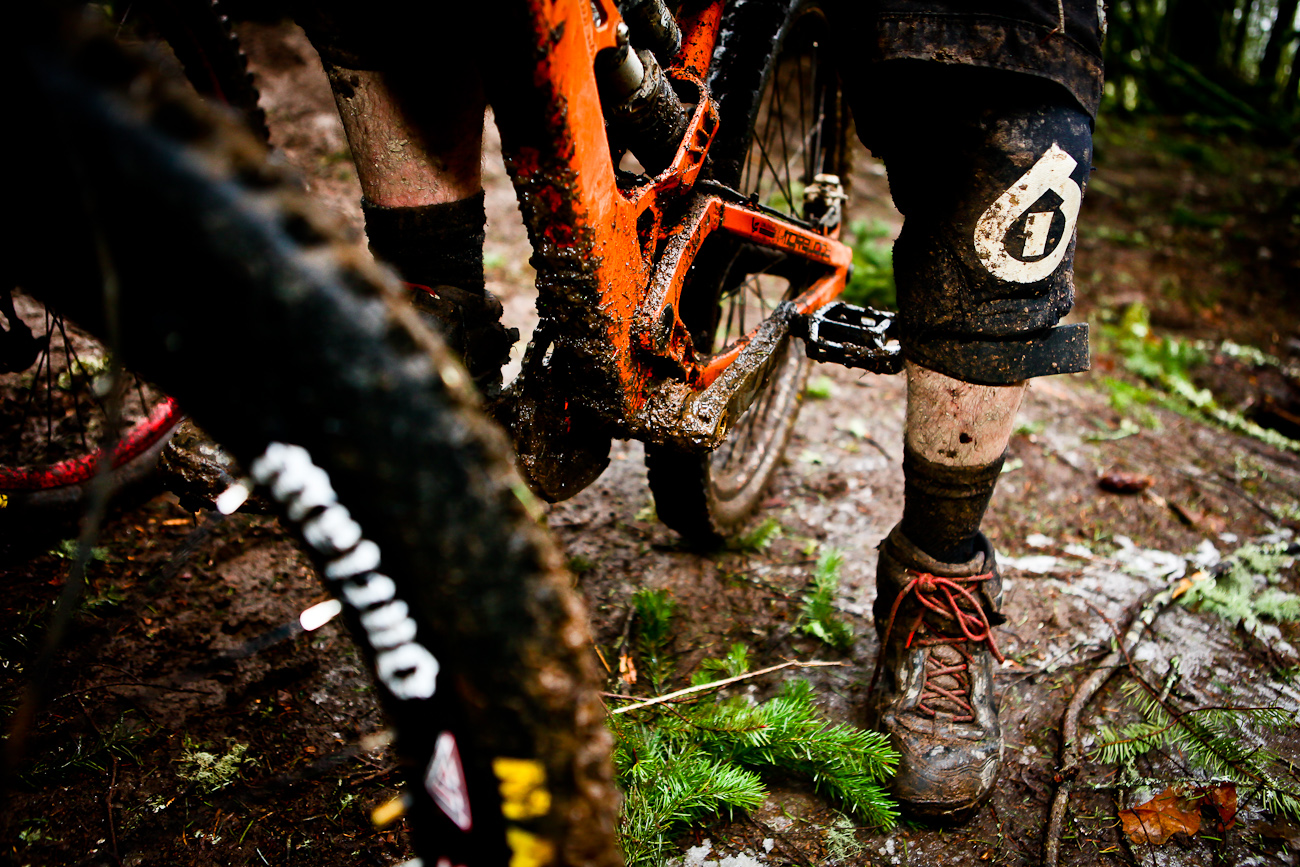 Legs of a mountain bike cyclist covered in sweat and mud. Their bright orange bike is also muddy. They are on a trail with pine trees and ferns.