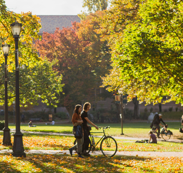 Students walk through the middle of campus in fall. A mix of red, green and yellow leaves are seen on the trees and the ground.