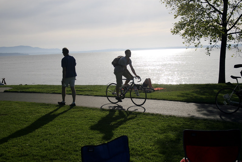 A student rides a bicycle through a park. The cyclist is backlit by the sunset over Bellingham Bay.