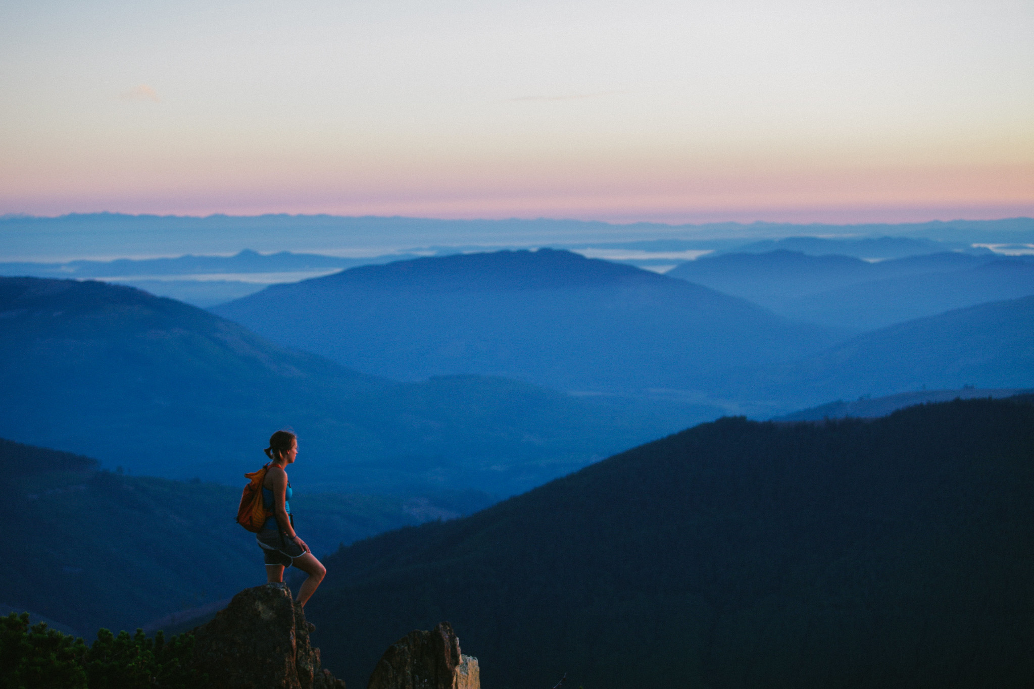 One hiker stands on top of a peak. From her viewpoint, we can see rolling hills all the way to the horizon. The sun is either just setting or just rising.