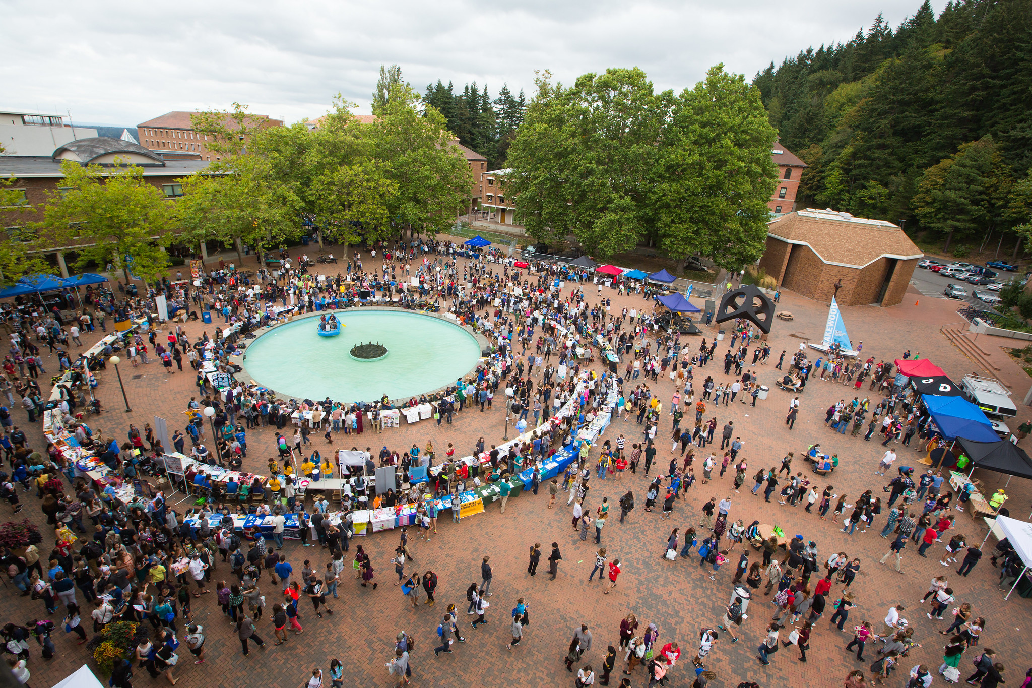 An aerial view of Red square, a brick courtyard lined with trees and buildings. There is a round fountain in the middle of the square, and two rings of tables and booths are set up around the fountain because an info fair is happening. There are hundreds of students interacting with each other and the tables.