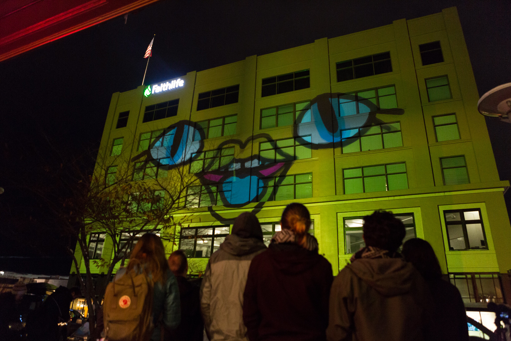 An animation is projected onto the side of the Flatiron building using a projector. The projected art makes the whole building look like a yellow cartoon face. A crowd of people watches the show.