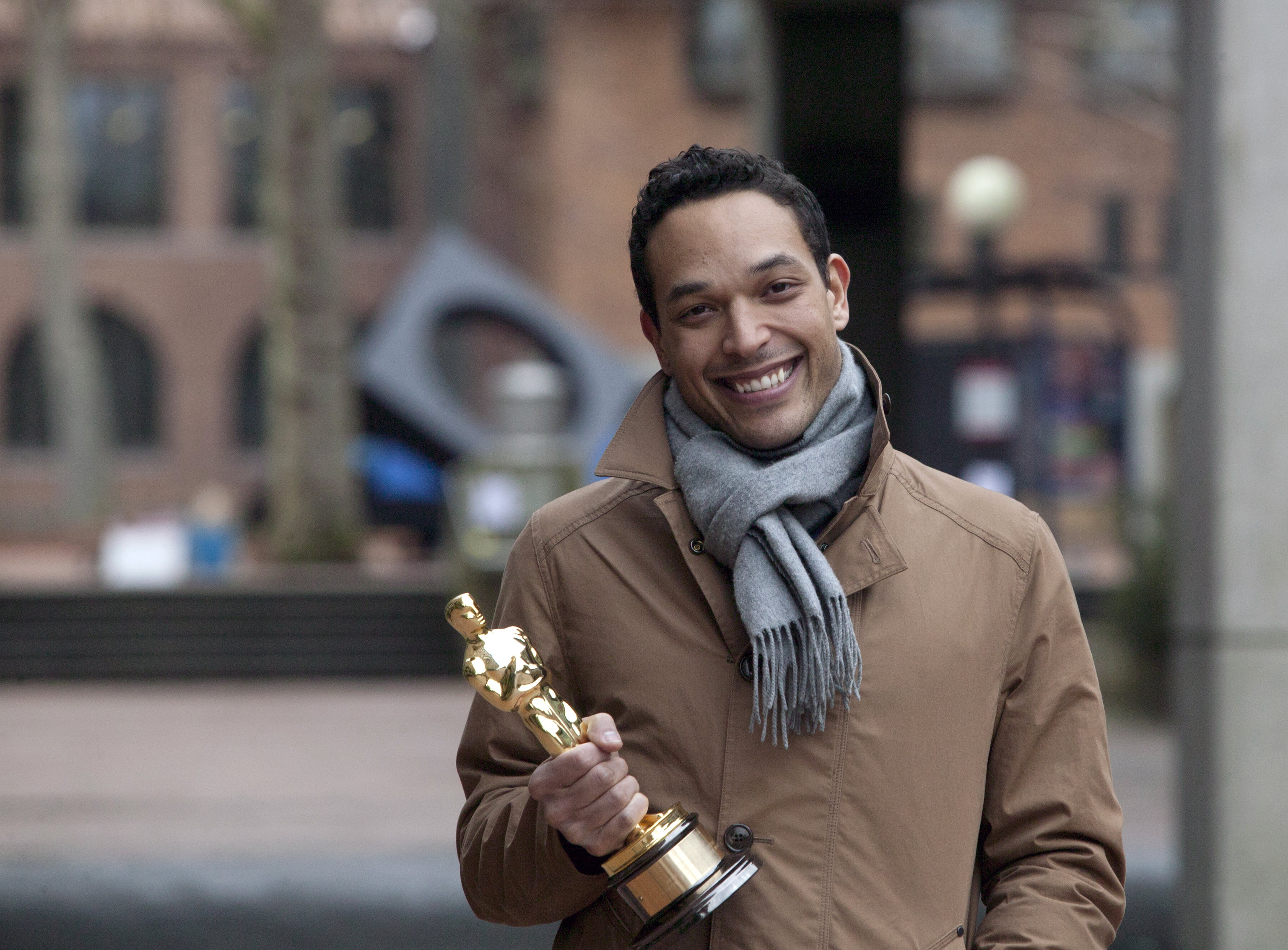 TJ is wearing a tan jacket and a thick scarf. He is smiling and holding his Oscar award. Behind him, Sky Viewing Sculpture and Miller Hall are visible.