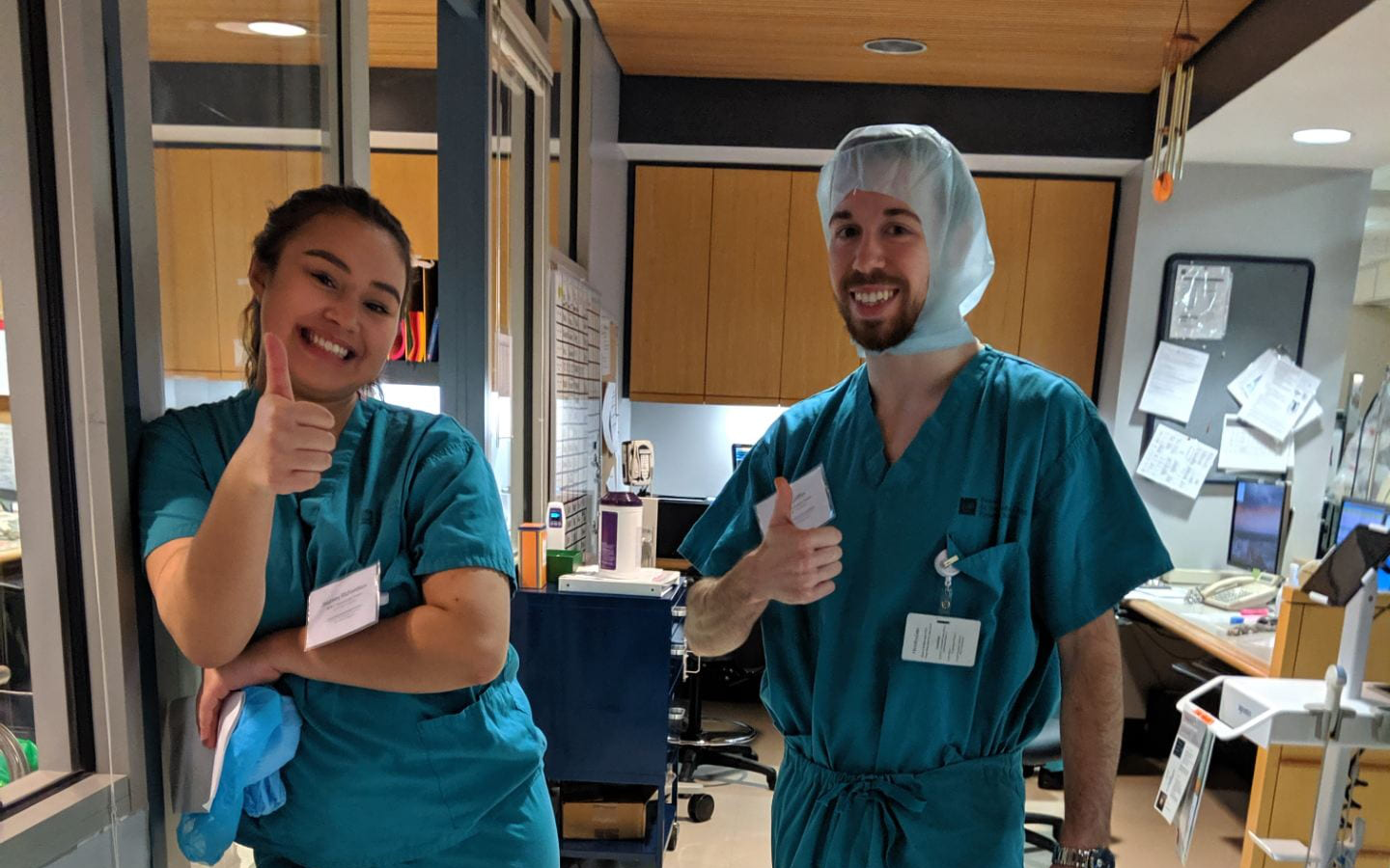 Students Journey and Kyle are wearing teal scrubs and giving the camera a thumbs up. They are in a medical setting at their behavioral neuroscience internship.