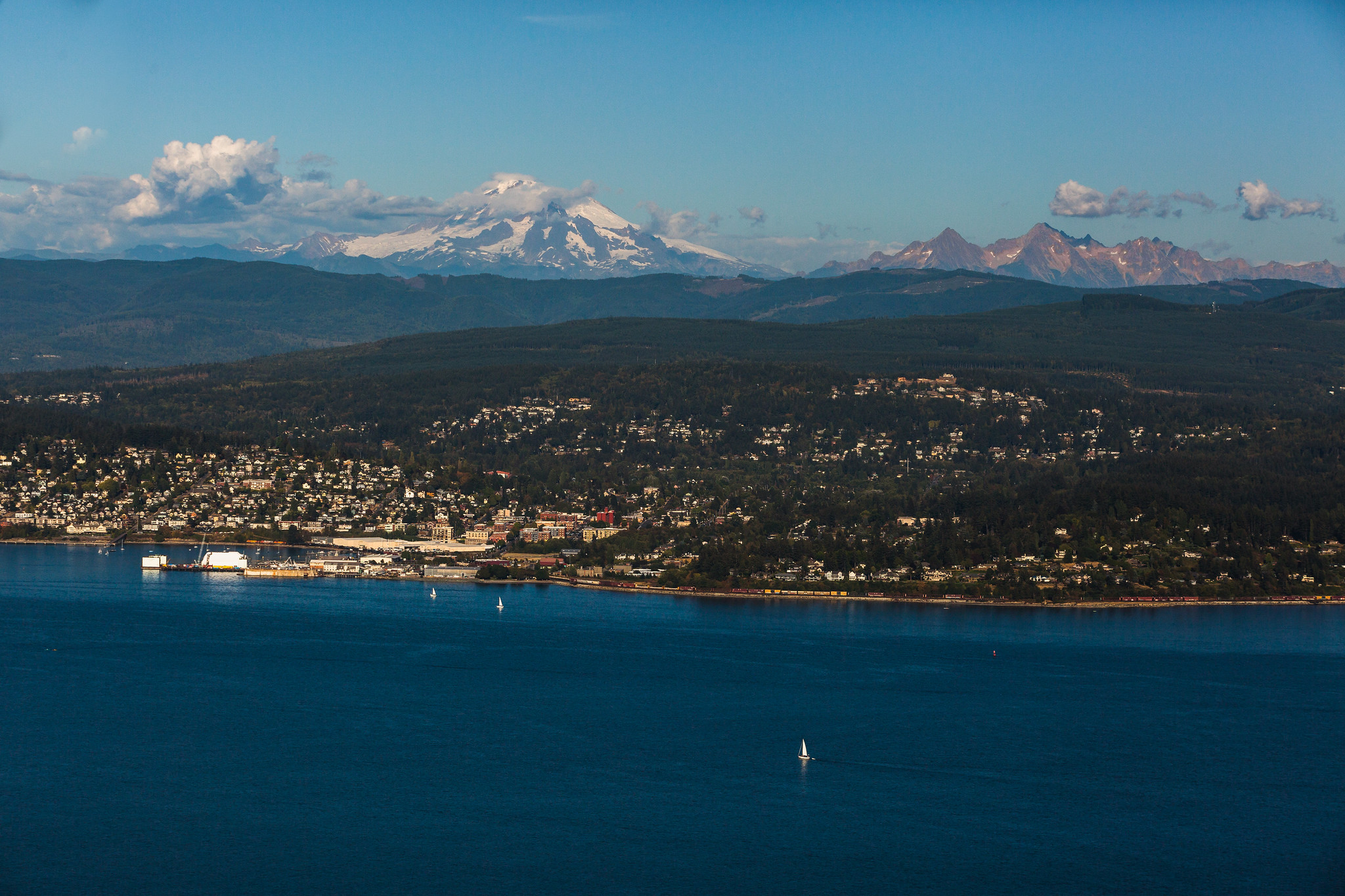An aerial view of Bellingham on a sunny day. Mount Baker is visible in the background, and the city of Bellingham and Western's campus are visible. Bellingham Bay fills the lower third of the photo.