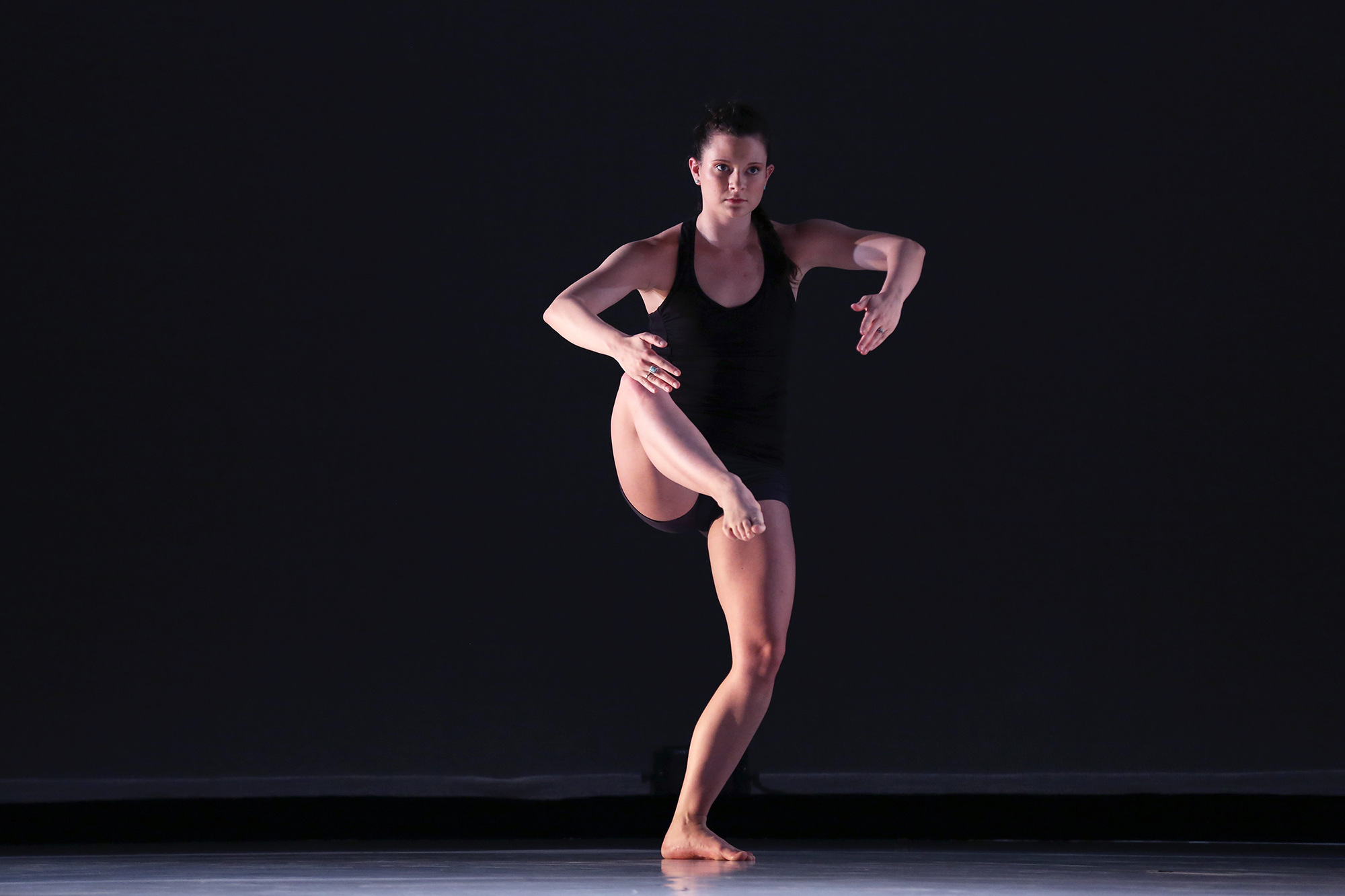 Brynne is dancing with her right leg lifted and bent, and her arms in front of her. She is wearing a black leotard and standing in front of a black backdrop. 