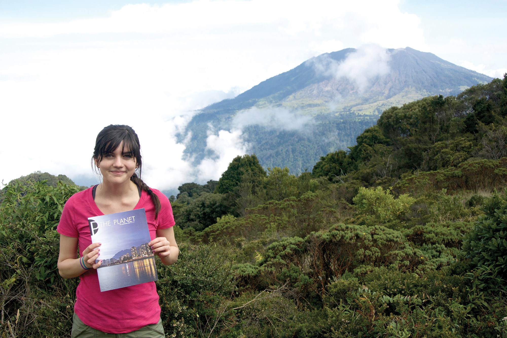 Yvonne is standing in a thick, green jungle holding a copy of The Planet magazine. A large hill or mountain is in the background, partially obscured by a low cloud.
