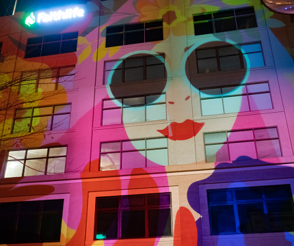 An illustration of a woman with pink hair, round sunglasses, and bright red lips is projected onto the side of the Flatiron building. The illustration covers several floors of closed windows.