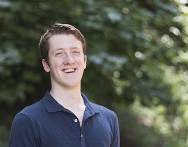 Evan is smiling and wearing a blue polo shirt. He is outdoors and there is sunlit greenery behind him.
