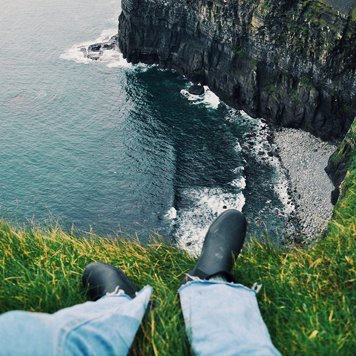 A student is sitting on a grassy slope with their feet dangling over the edge. The ocean is visible far below, and there are large cliffs in the background.