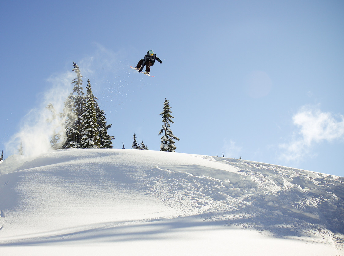 A snowboarder is in a mid-air jump, trailing powder behind them against the blue sky.