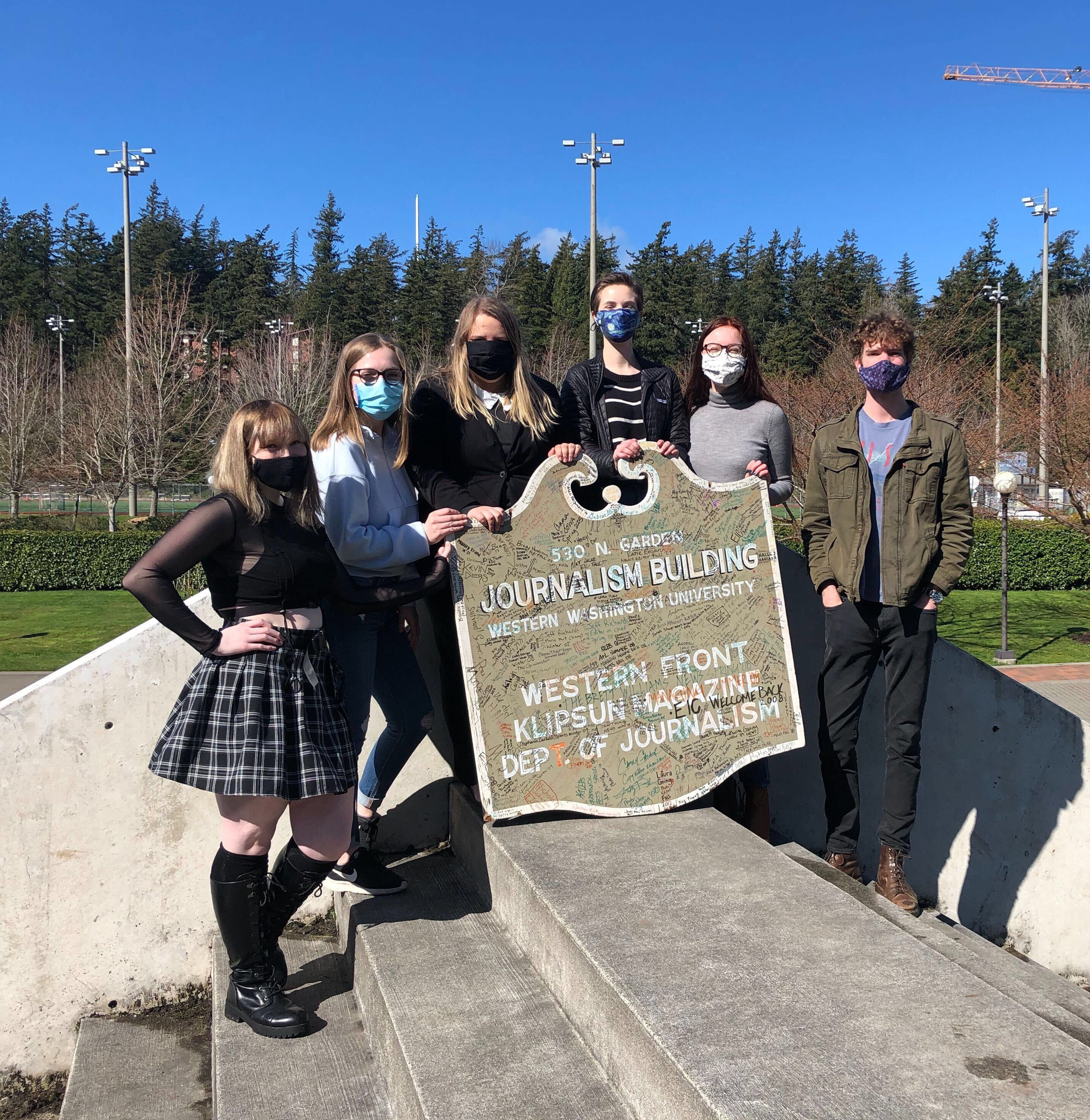 Six students are standing on top of the Stadium Piece sculpture, all facing the photographer. They are holding a plaque that says "Department of Journalism Western Washington University" and has hundreds of signatures on it.