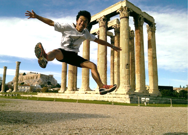 A student is in a mid-air jump with his arms and legs stretched out. Behind him is an ancient structure built of ornate Greek columns called the Temple of Zeus. Additional columns and the ancient Parthenon are visible in the background. 