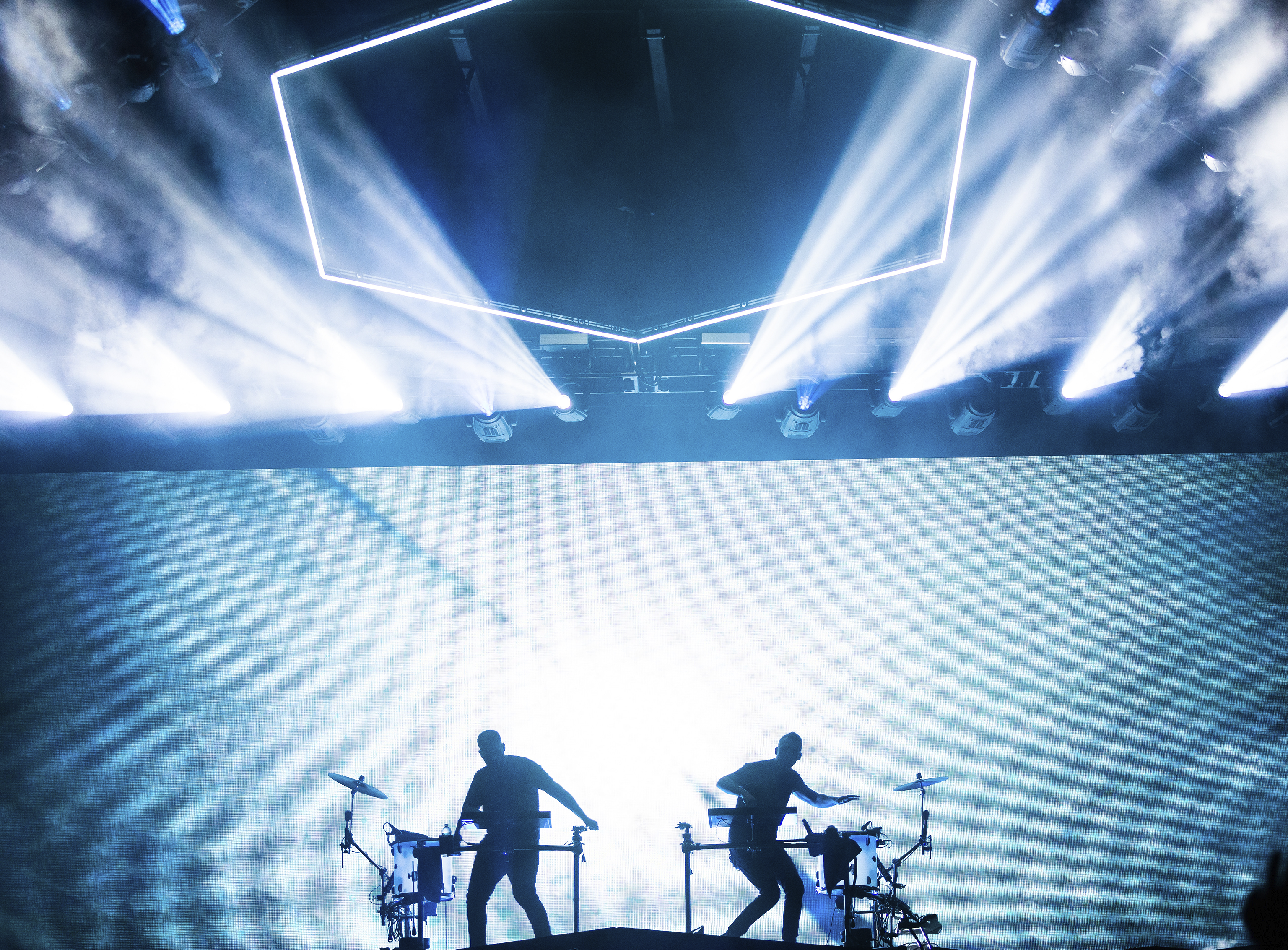 Two musicians in the band Odesza are on stage at a concert, backlit by a giant glowing blue screen. Each man is playing half of a drum kit. Above them, a glowing hexagon is suspended in the air and spotlights are lighting up the night sky. 