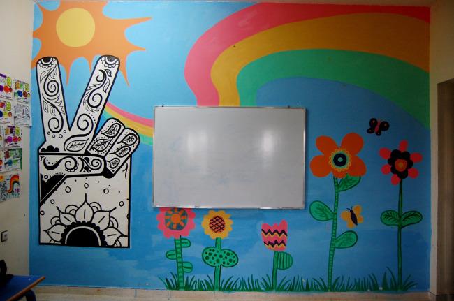 A mural painted on an interior wall. The background is sky blue, with a sun, rainbow and flowers obviously painted by children. There is also a stylized hand making a peace symbol on the wall.