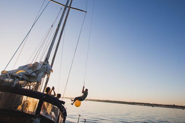A medium-sized boat is on Bellingham Bay at sunset, and the water is calm. A person is swinging on a buoy or ball that is suspended from the mast, they are swinging far over the side of the ship above the water. 