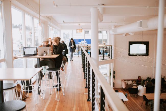 Students are looking at laptops and notebooks in a cafe. The cafe has two floors, so we see students on the upper balcony as well as on the floor beneath them. A wall of tall windows lets natural light into the very white room.