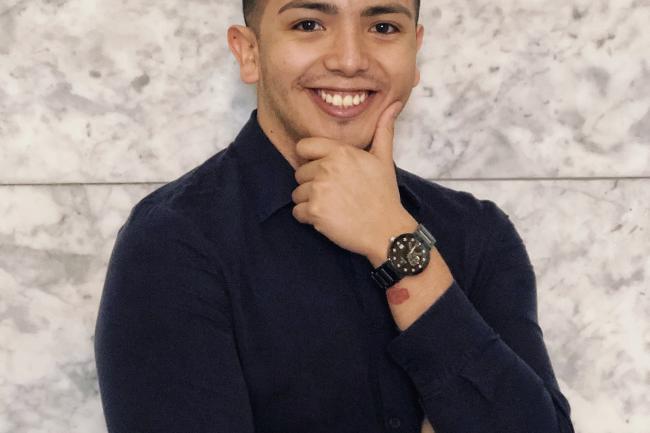 Ranulfo is smiling and has one hand held to his chin. He is wearing a dark button down shirt and a nice watch.