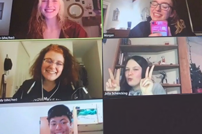 Five students are in a zoom meeting together. They are all smiling, one is wearing a paper crown, one is flashing peace signs with their fingers, and another is using their phone to take a group photo.