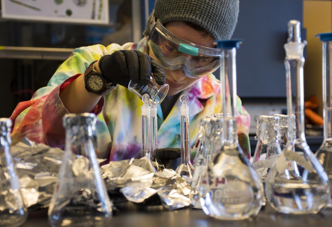 A student in a tie-dyed lab coat uses laboratory glasswear. 