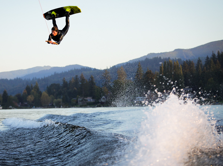 A wakeboarder is upside down in mid air. There is a large wave and beneath them, and there are scenic hills and trees far in the distance behind them.