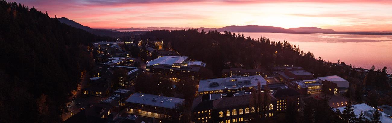 Campus at sunset in the wintertime. The sky, islands, and bay are pale yellow, peach, and lavender. Campus is tucked into the dark shadow of Sehome Hill. Campus is still active and many lights glow gold.