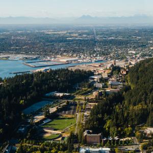 An aerial view of WWU campus in the foreground and Downtown Bellingham in the background. To the left is the bay and far in the back is a ridge of mountains on the skyline, peaking through the mid-day haze.