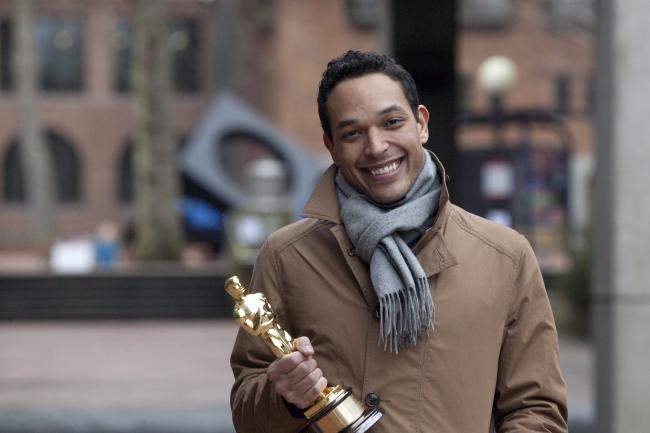 TJ is wearing a tan jacket and a thick scarf. He is smiling and holding his Oscar award. Behind him, Sky Viewing Sculpture and Miller Hall are visible.