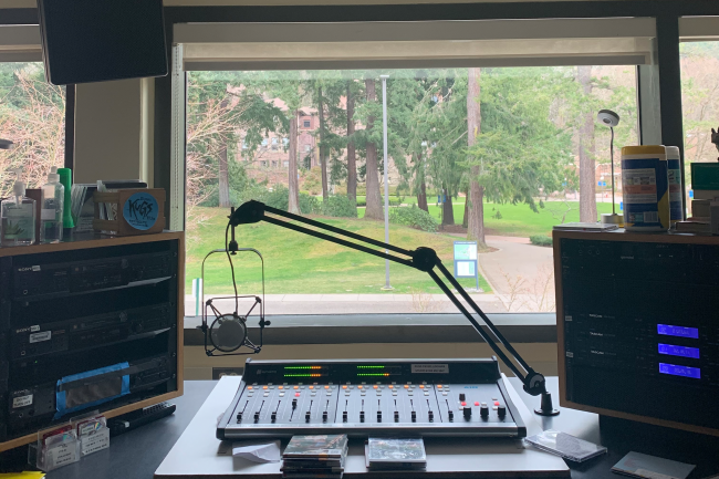 This is the view KUGS disc jockeys see from their station. There is a soundboard and microphone in the foreground, in front of a window. Out the window, grass and trees are visible.