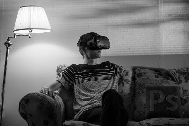 A moody, noir black and white image of Zach, who is alone on a couch wearing Virtual Reality goggles. The room is illuminated by a solitary lamp.