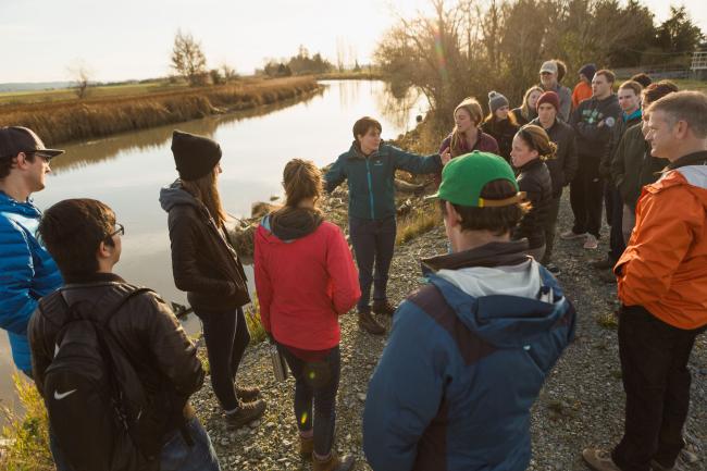 A class stands watching their professor teach next to the Skagit River.